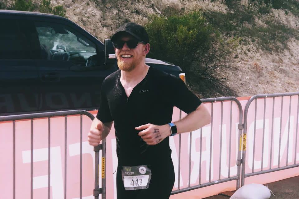 Craig mid-stride at the Wildflower Triathlon, in a black tri suit, grinning past the course barriers.