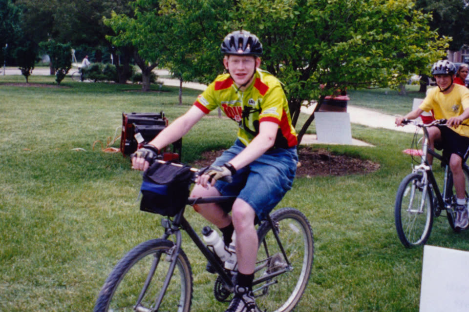 Craig at 13, in a bright cycling jersey and helmet, riding a mountain bike across a park lawn at the Tour de Cure.