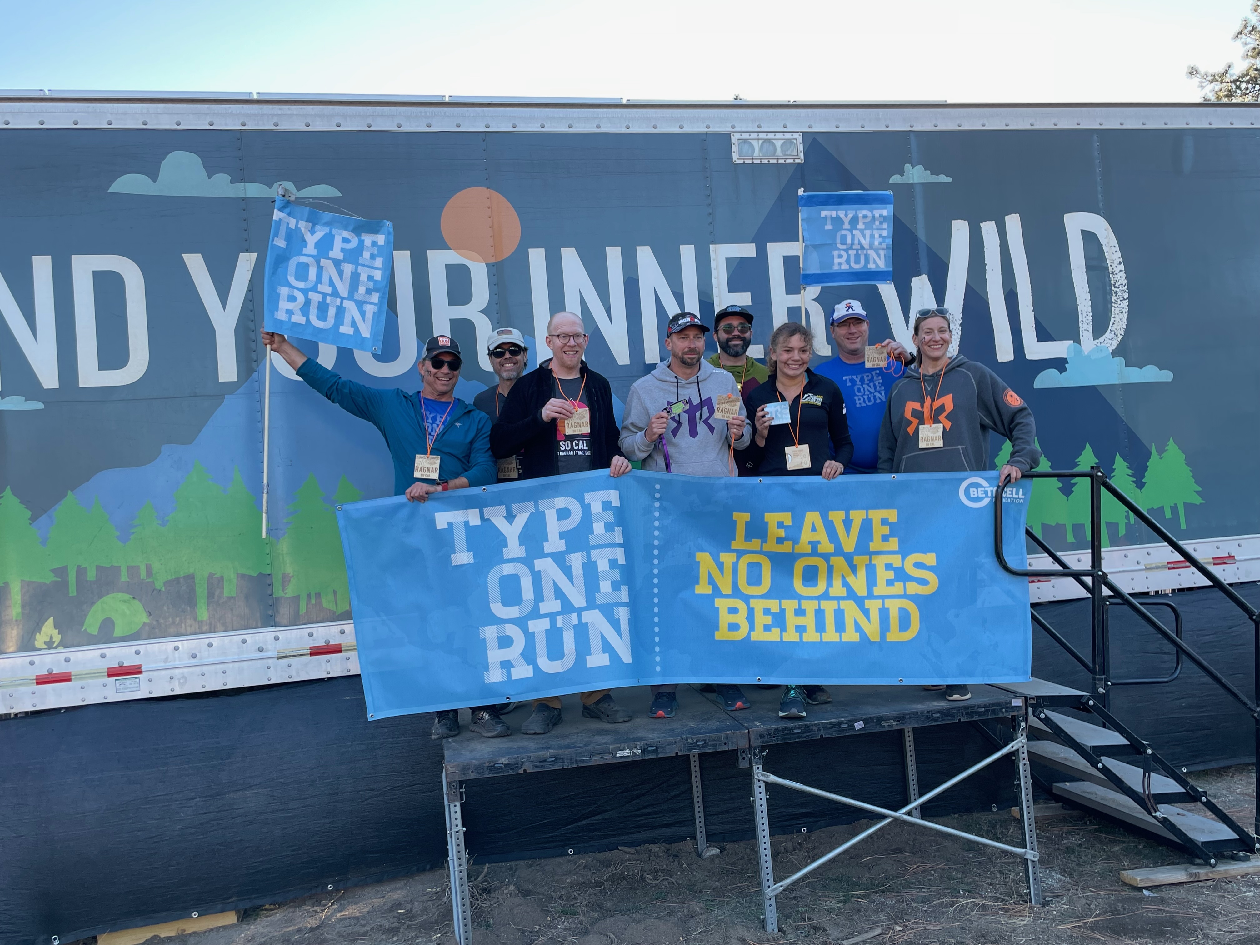 Eight runners holding banners and medals posing together in front of a branded event trailer after completing a race
