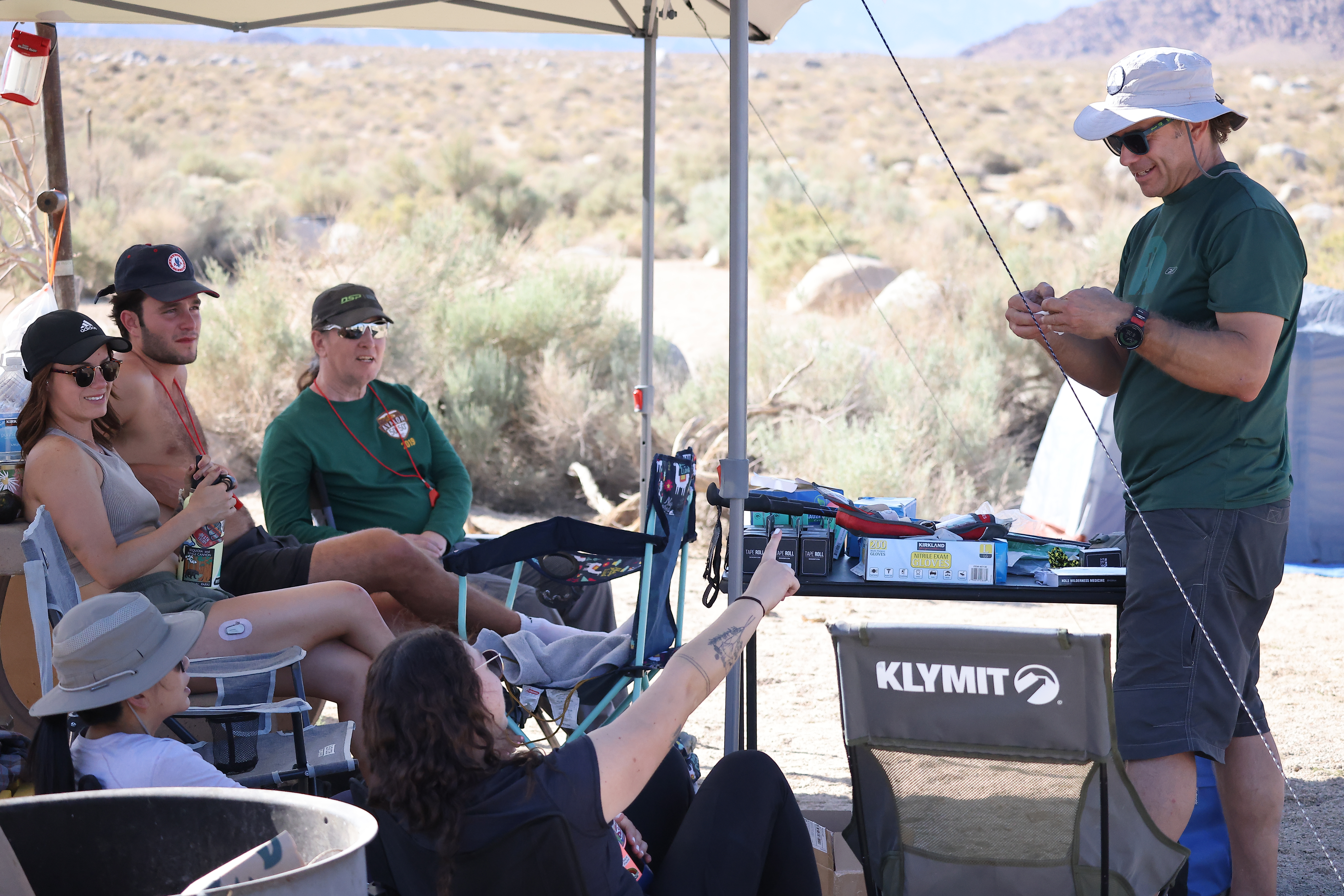 Group of five people relaxing under a canopy shade structure in a desert campsite, one person rigging a fishing rod