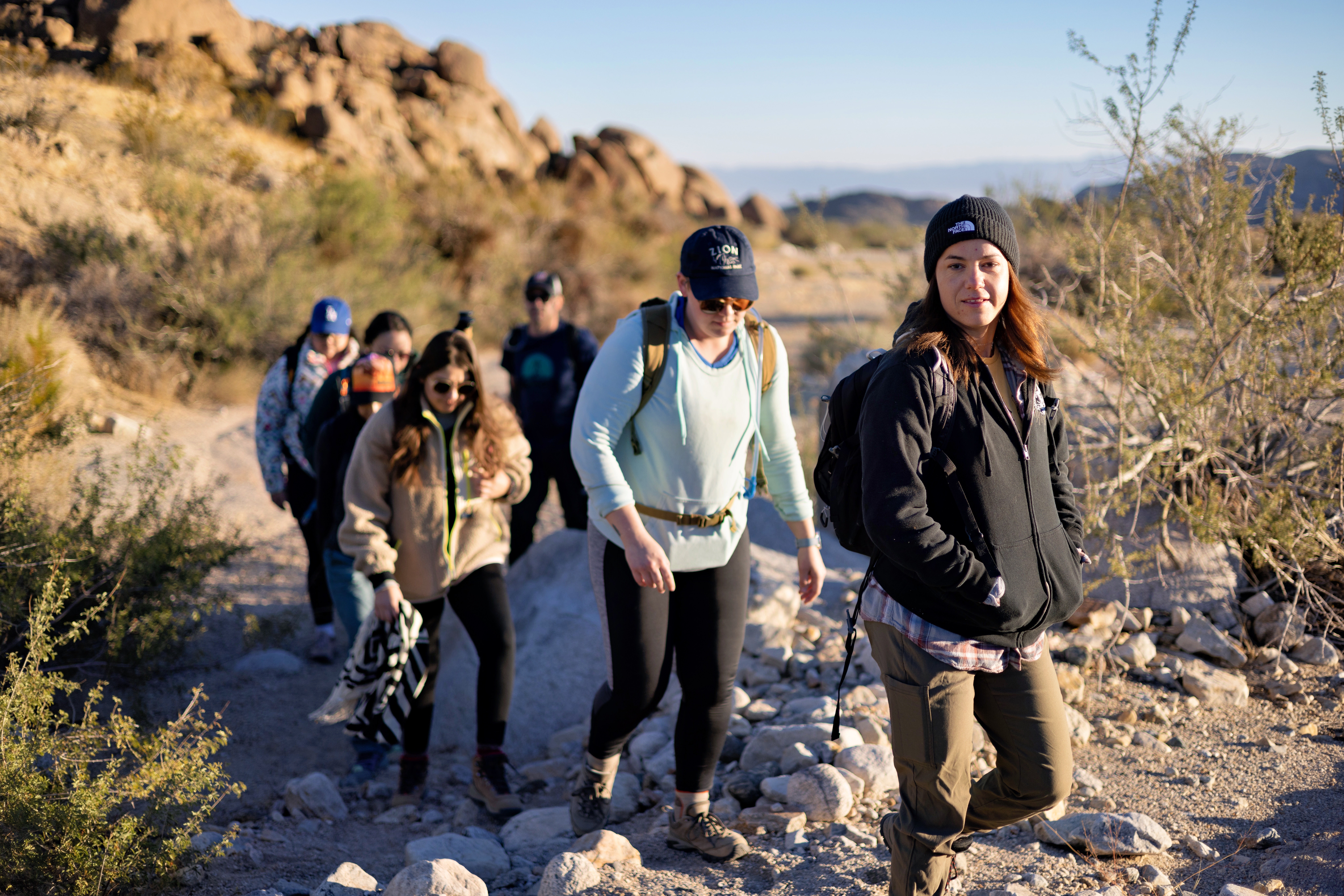A group of six hikers walking single file along a rocky desert trail with large boulder formations in the background