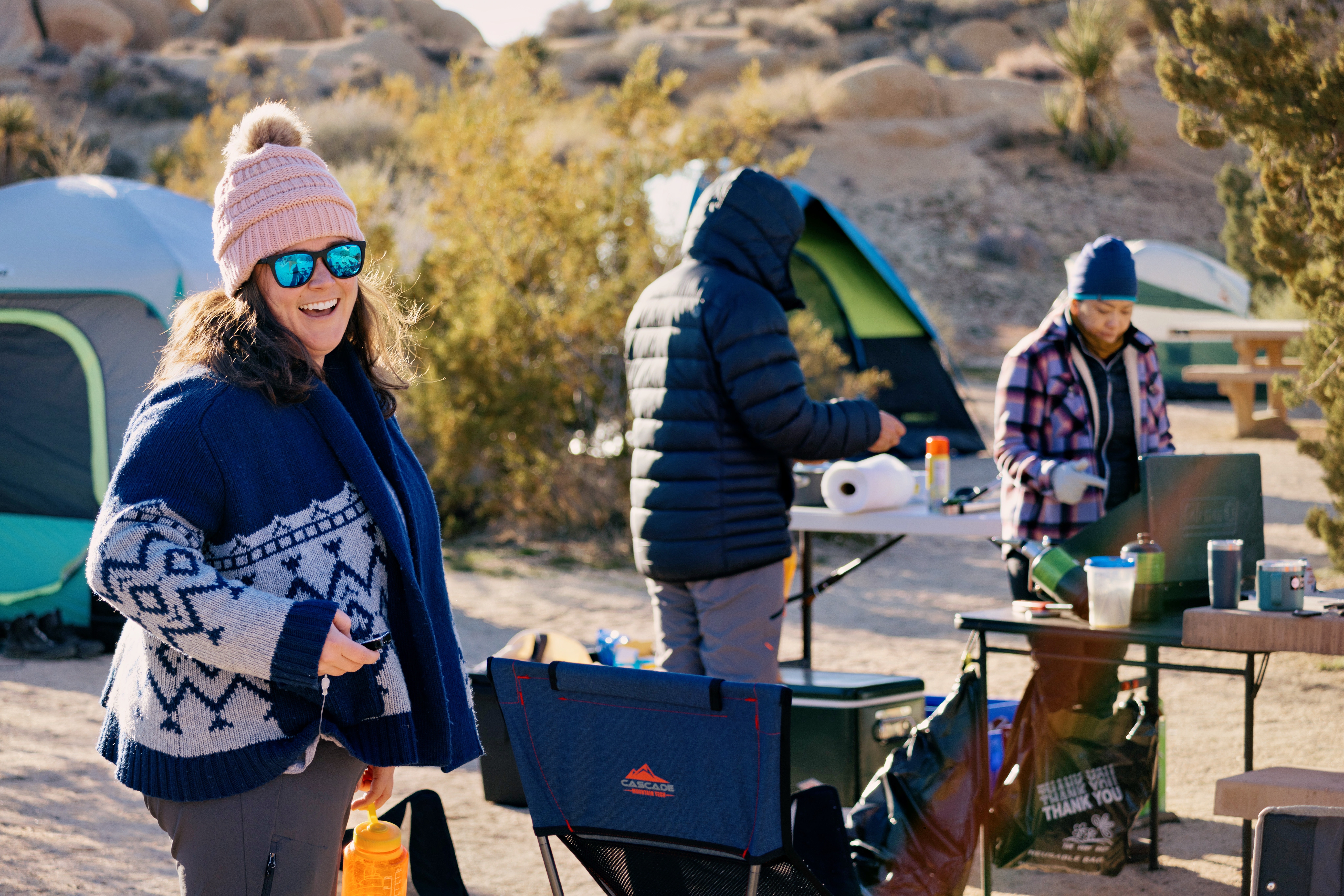 A woman in a knit sweater and pink beanie smiling at a desert campsite while two others prepare food at a camp table