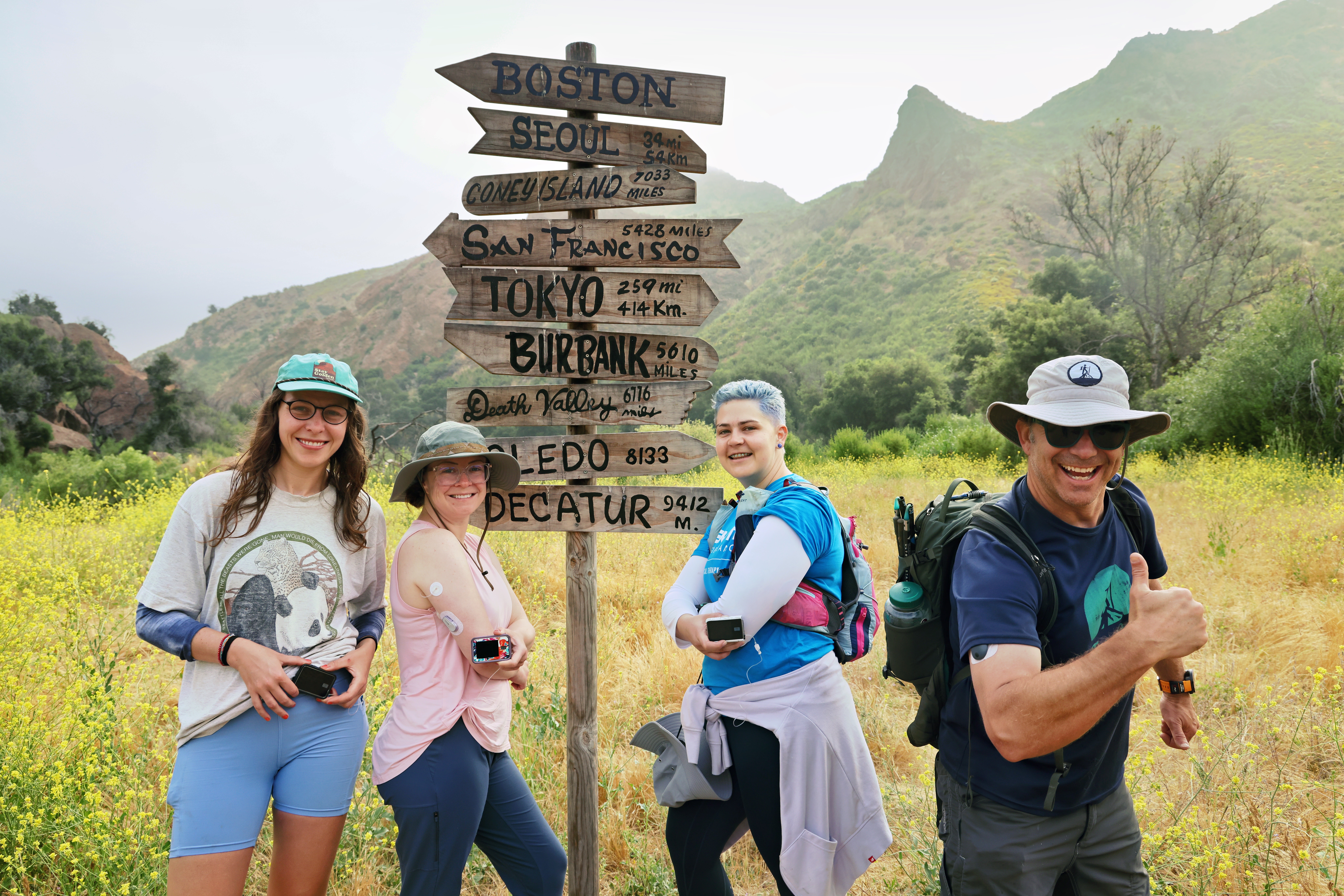 Four smiling hikers with backpacks posing next to a rustic wooden directional signpost in a wildflower meadow